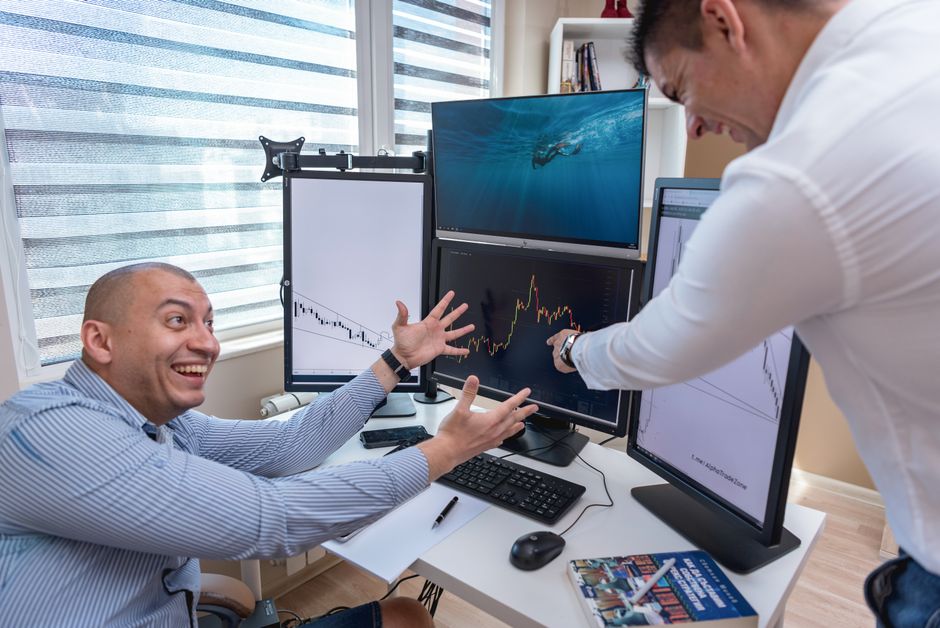 Overcome trading fomo — Two businessmen enthusiastically analyzing market charts on computer monitors in a modern office.
